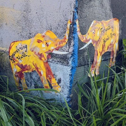 Photograph of a pair of bright yellow painted elephants, one on each side of a corner in a concrete wall. One elephant has the words 'remembering hurts...' written on it, while hte other has the words 'forgetting hurts more'.