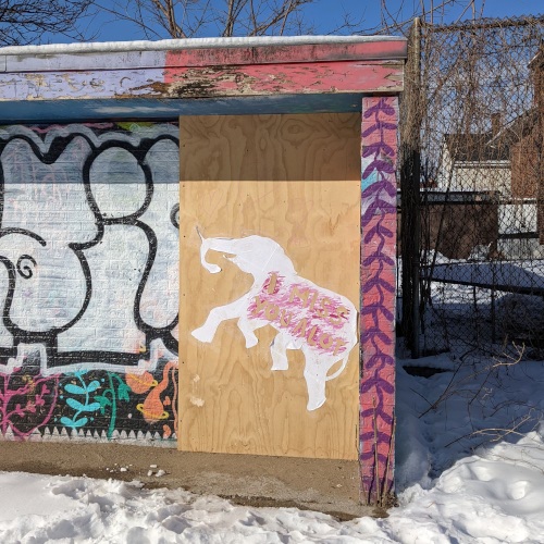 Photograph of a small graffiti-covered building, with a white elephant pasted on top of some plywood. some not-very-legible text reads 'I miss you a lot'.