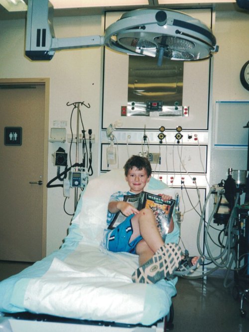 Photograph of a tousle-haired child, maybe eight years old, sitting in a hospital bed with sneakers on, smiling at the camera. He is reading a large hardcover Warhammer book.