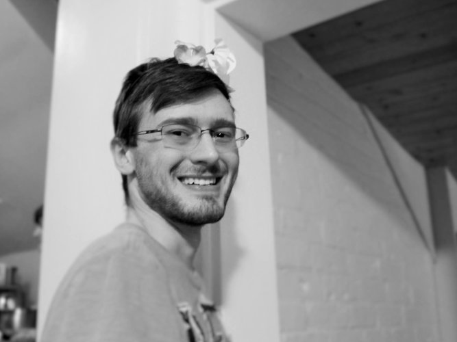 Soft black and white photograph of Zachary Jacobi looking at the camera, with a flower crown in their hair. They are smiling wide.