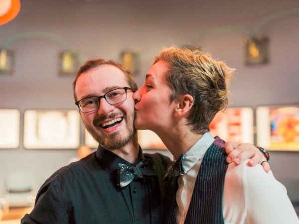 Photograph of Zachary Jacobi and I, wearing matching outfits. I am kissing Zach's cheek and they are grinning.