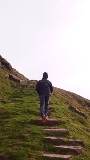 Photograph of Zachary Jacobi climbing a set of stone stairs on the side of a hill in Wales. The hill is a fae green and the sky is a pure cloud white.